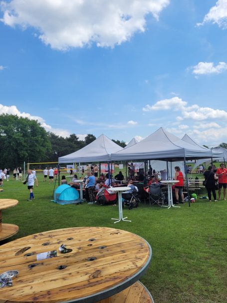 Menschen sitzen unter Zelten auf einer Wiese, während im Hintergrund Fußball gespielt wird.