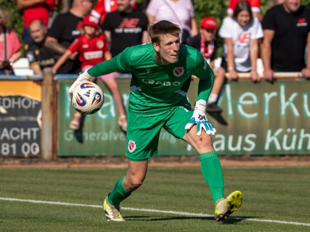 Torwart im grünen Trikot fängt den Ball, Fans jubeln im Hintergrund.
