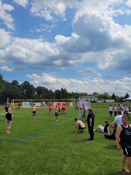 Volleyballspiel auf einem grünen Feld, viele Menschen und bewölkter Himmel im Hintergrund.