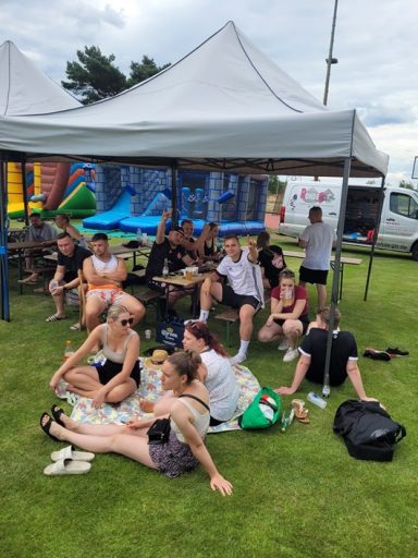 Jugendclub Dissenchen Gruppe von jungen Menschen sitzt unter Zelten auf einer Wiese, mit Spielgeräten im Hintergrund.