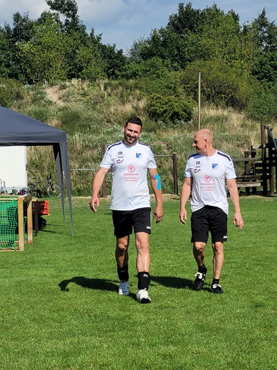 Zwei gut gelaunte Volleyballspieler im Trikot gehen auf einem grünen Sportplatz.
